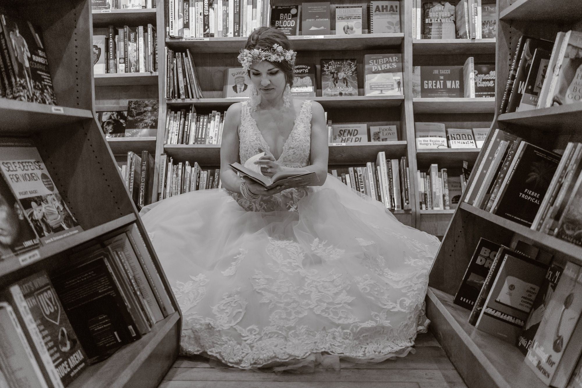 Black-and-white photograph of Leah reading in a wedding dress between bookstore shelves
